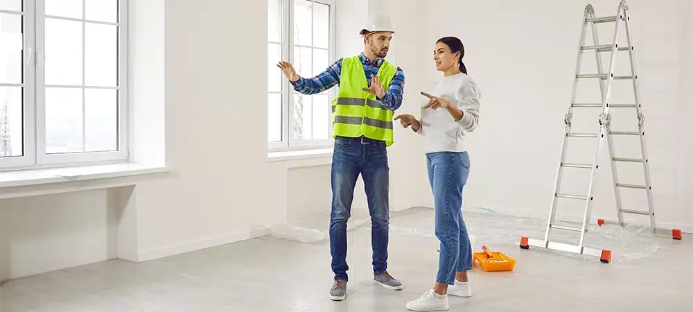 young woman homeowner discussing design project or repair of her new apartment with foreman standing in empty white room