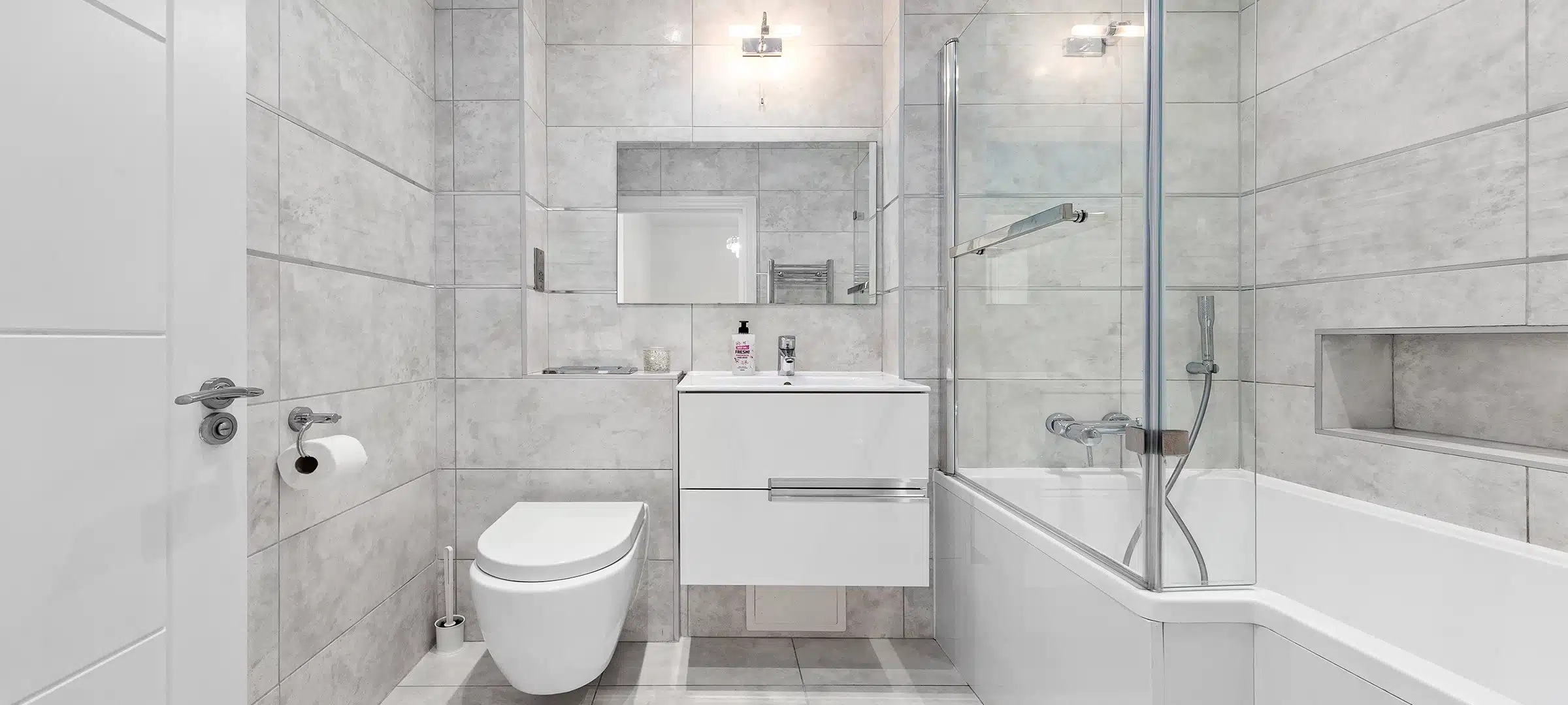 Sleek bathroom with large grey tiles, floating vanity, wall-hung toilet, shower-bath combo, glass screen, and recessed shelf with soft lighting