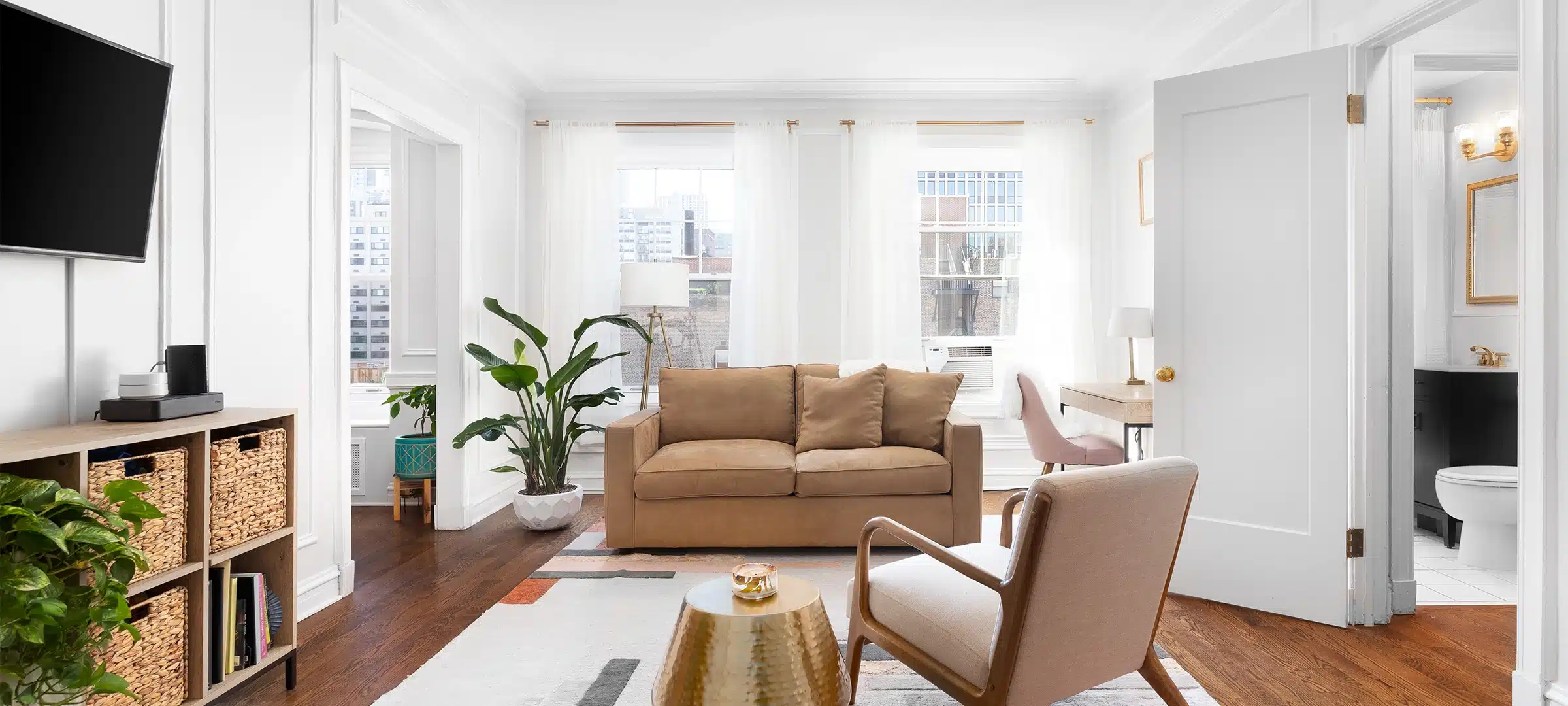 A bright, white living room in a downtown condo with cozy furniture, a television mounted on the wall, and a rug sitting on hardwood floors