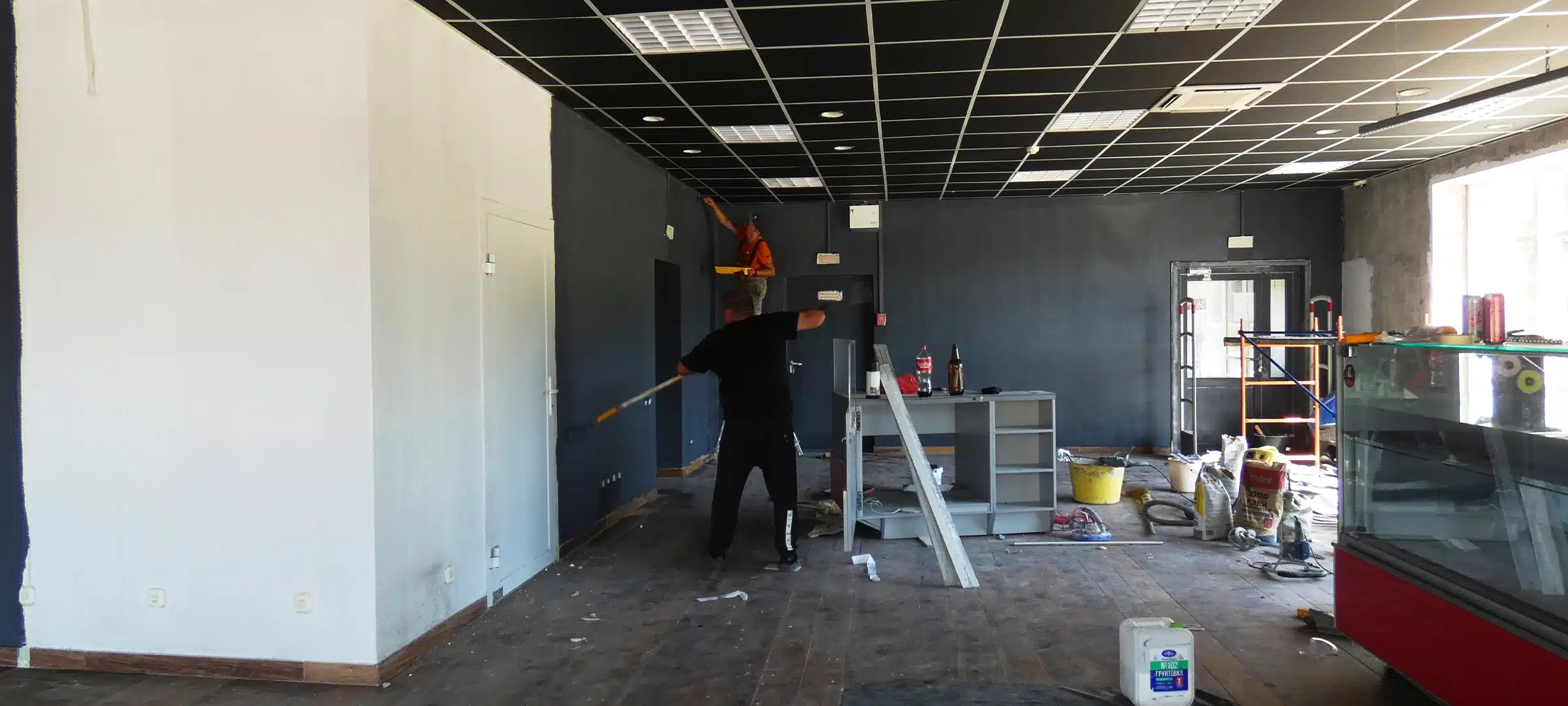 Workers are busy renovating a commercial space. One person is painting a wall, while another organizes tools and materials on the floor,