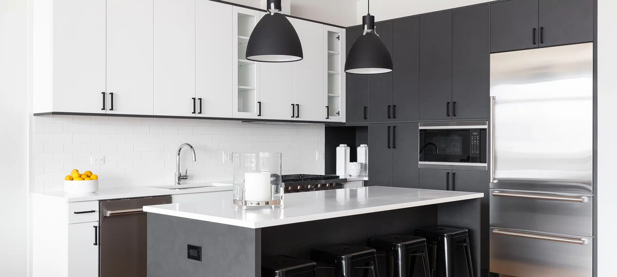 A modern kitchen with white and black cabinets, bar stools sitting at a large island, black pendant lights hanging over the marble countertop, and a subway tile backsplash. No brands or labels.