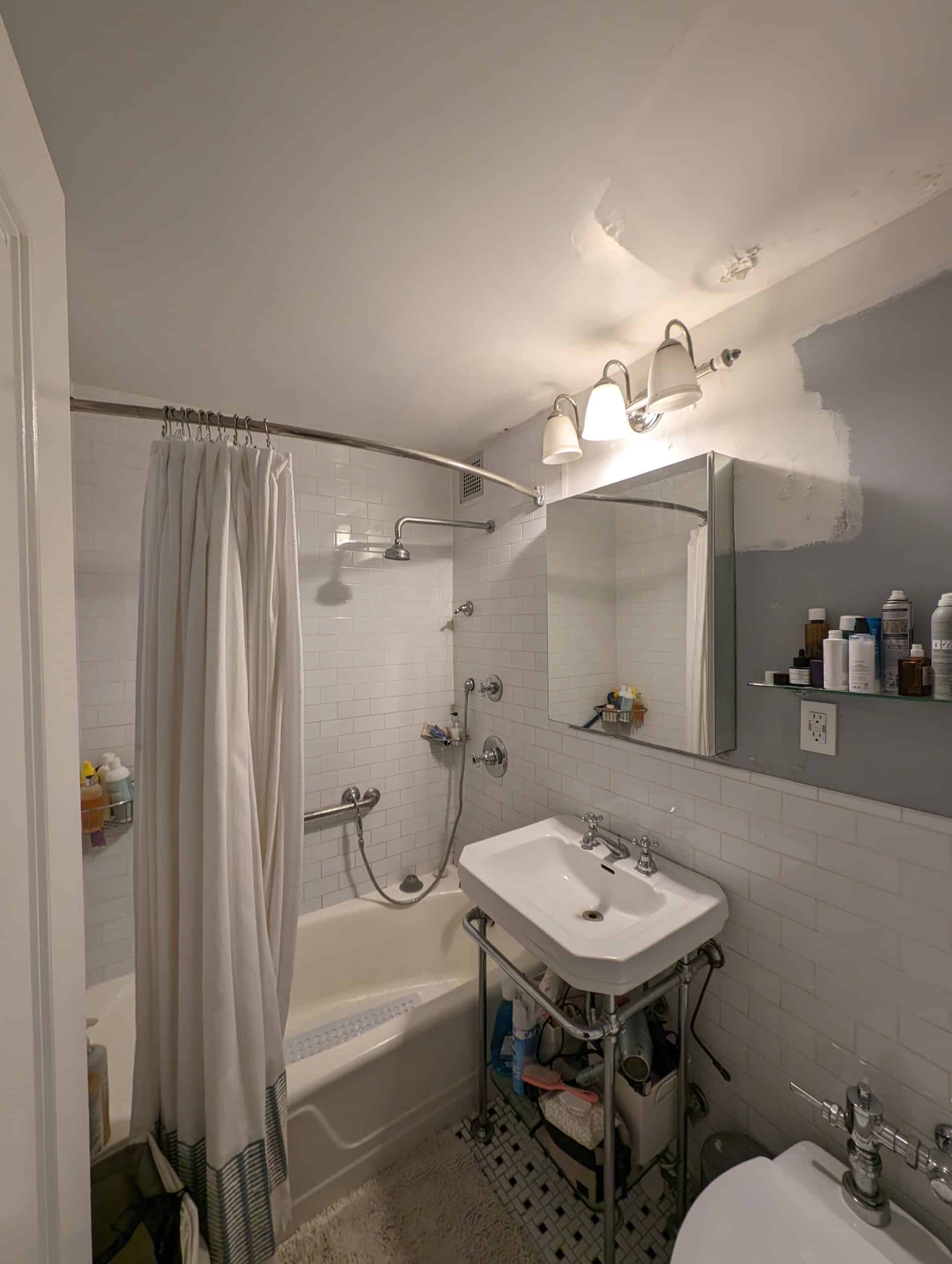 Before renovation: the second original bathroom at 230 East 79th Street with white wall tile, a wall-hung sink on exposed legs, a tub with a curtain and a mosaic floor