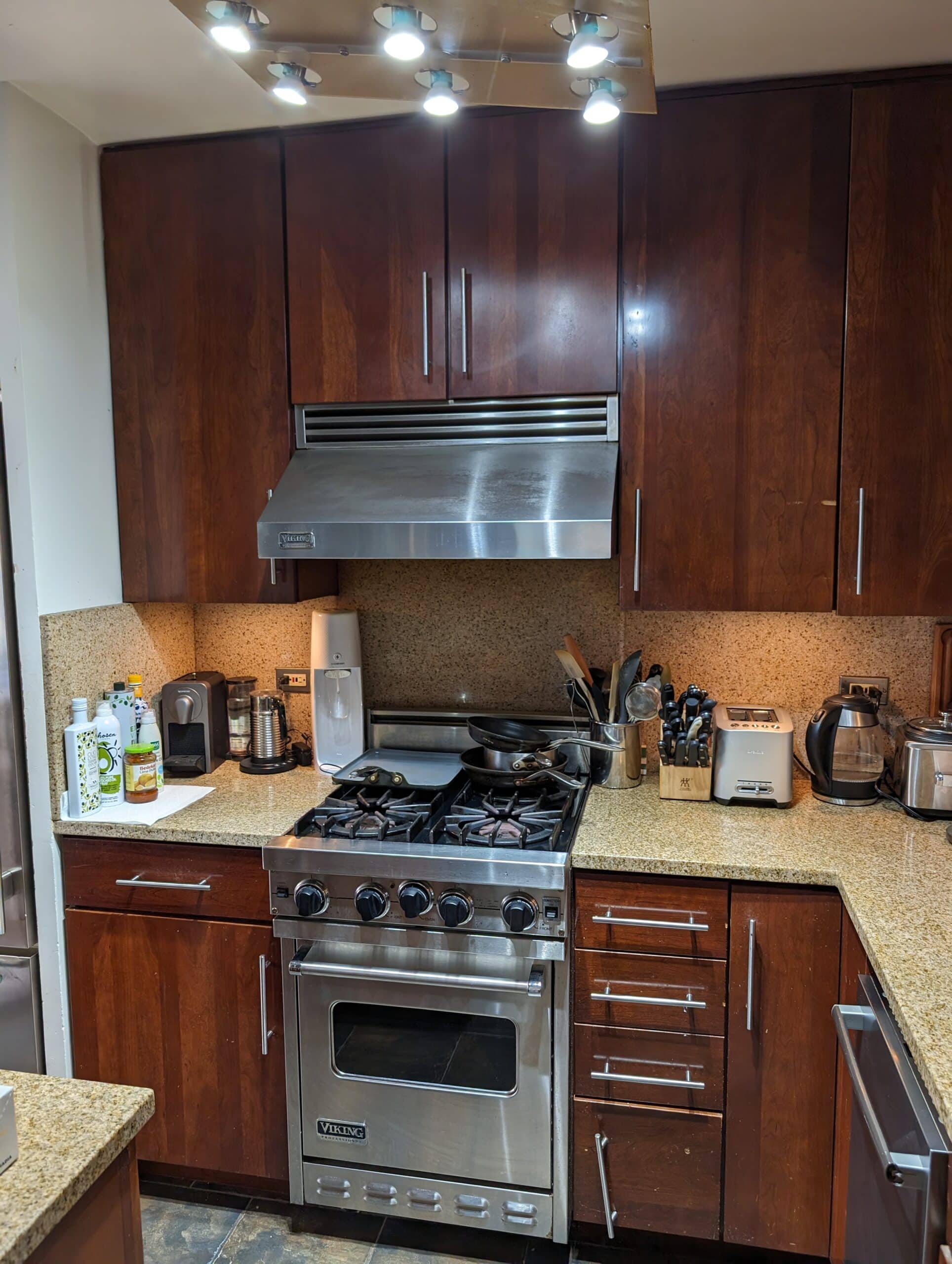 Before renovation: the original kitchen at 230 East 79th Street with dark cherry cabinets, a granite countertop, a tiled backsplash and a stainless Viking range under track lighting