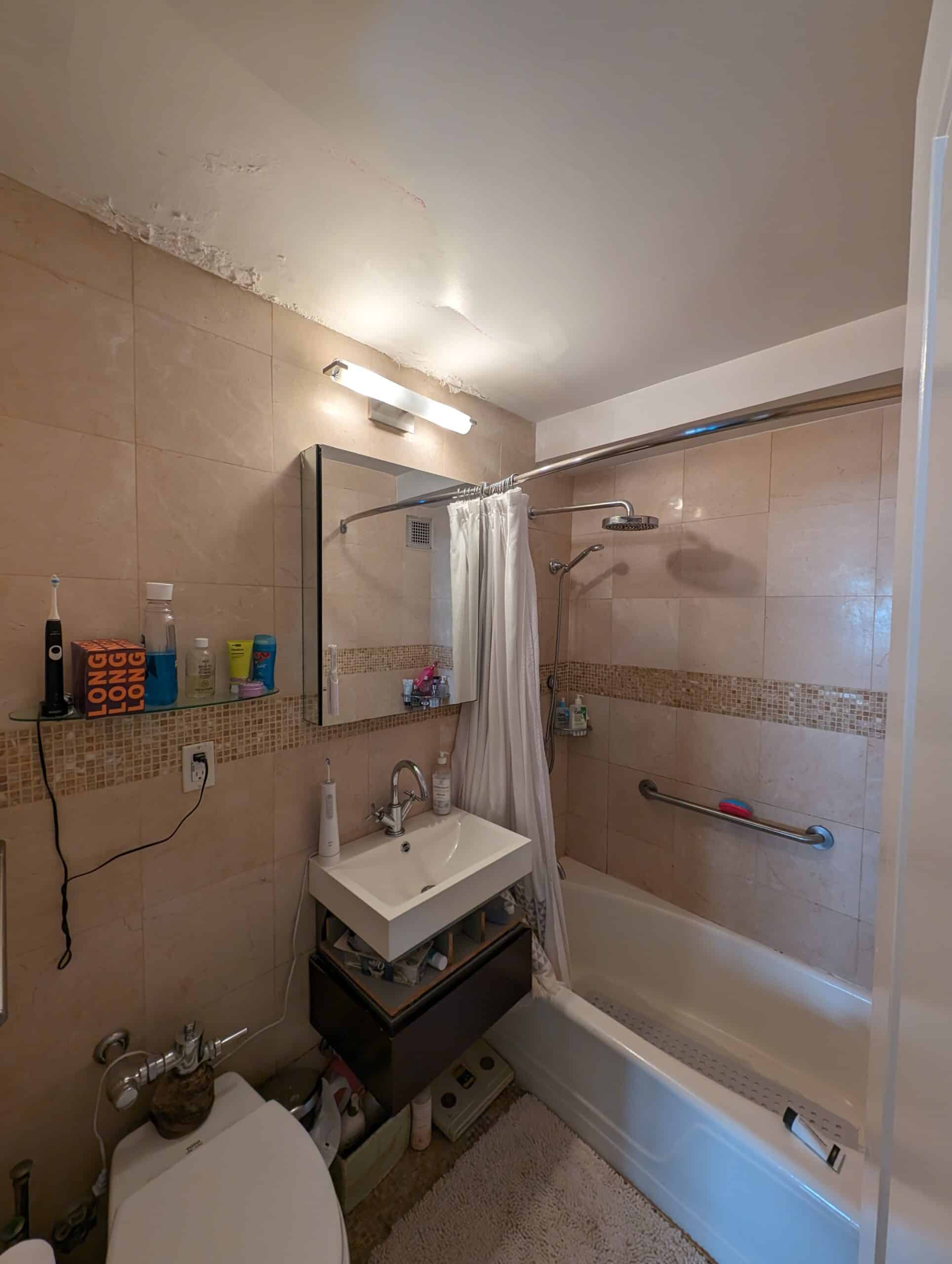 Before renovation: one of the original bathrooms at 230 East 79th Street in beige tile with a mosaic border, a wall-hung sink, a surface-mounted medicine cabinet and a tub behind a curtain