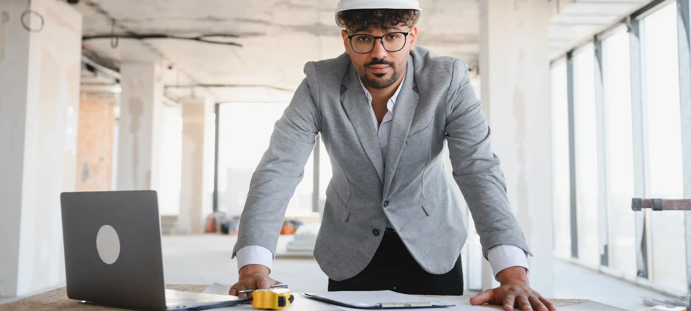 Arab architect wearing hardhat leaning on table with blueprints and laptop, overseeing construction project inside building