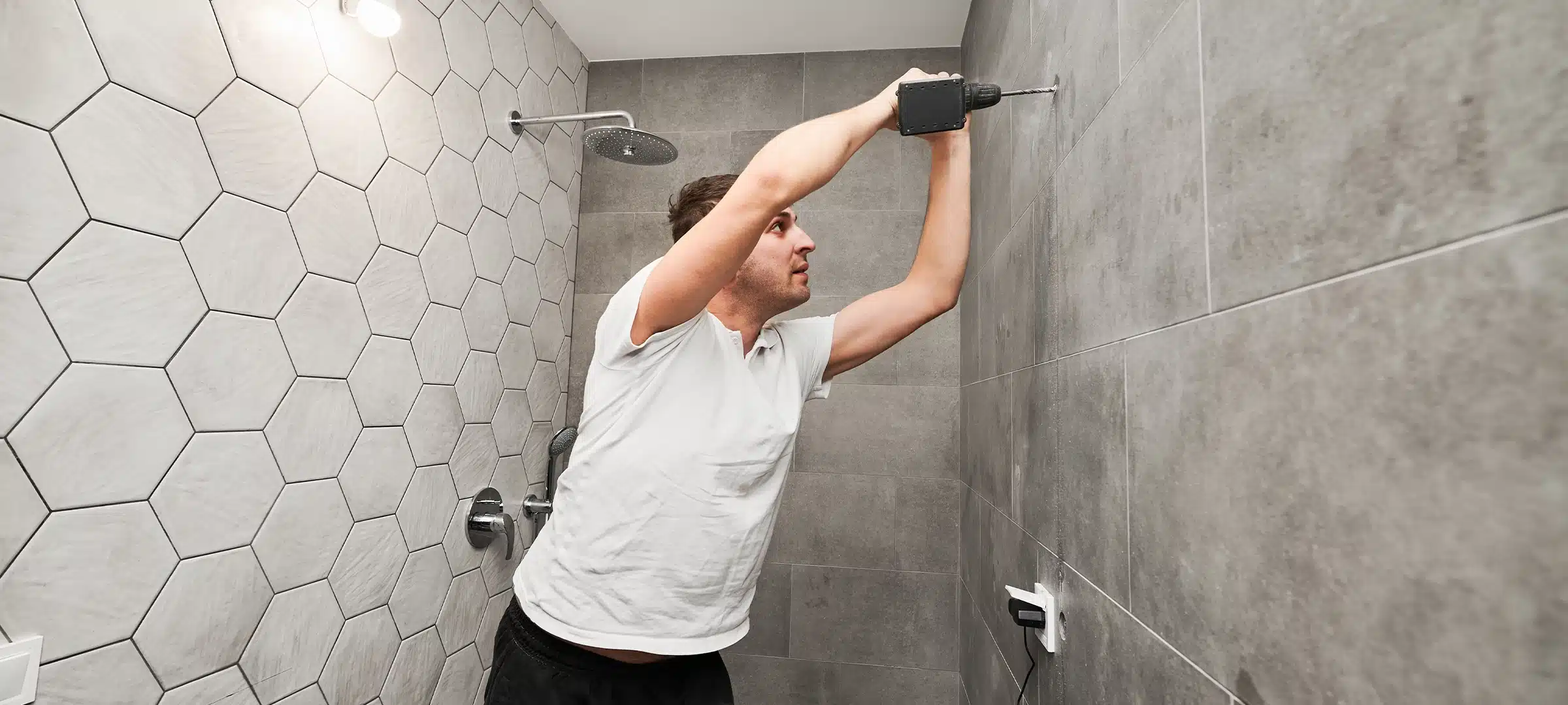 Side view of male worker using electric drilling machine while working on bathroom renovation. Man drilling wall with cordless power drill while installing shower system at home.