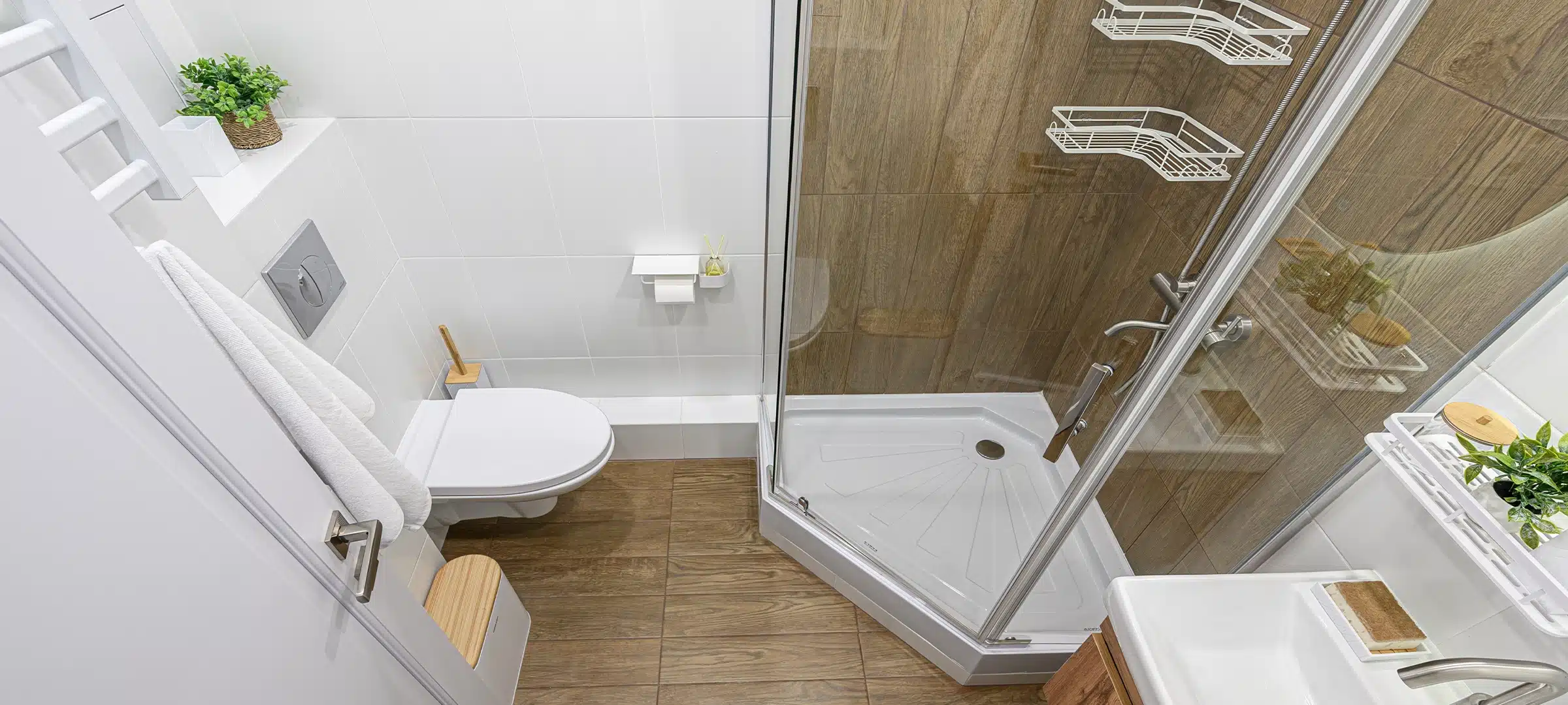 Modern bathroom with wood-look tile flooring, white tiles, shower stall, toilet, and vanity. Small plants add a fresh touch. Clean and efficient design
