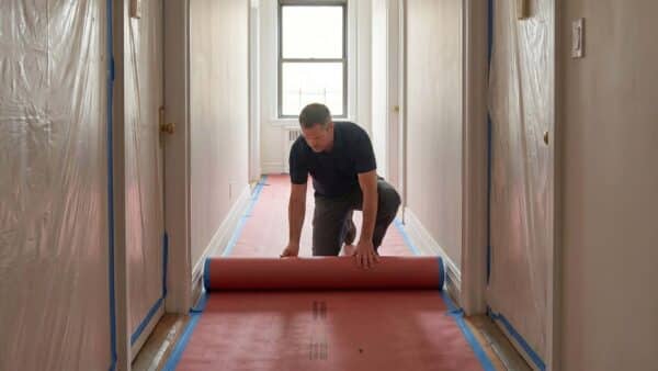 A male worker in a blue shirt is kneeling and rolling out a red protective runner, labeled "RAM Board," along a hallway floor in a residential building. The walls are covered with plastic sheeting and blue painter's tape, with a light fixture above.