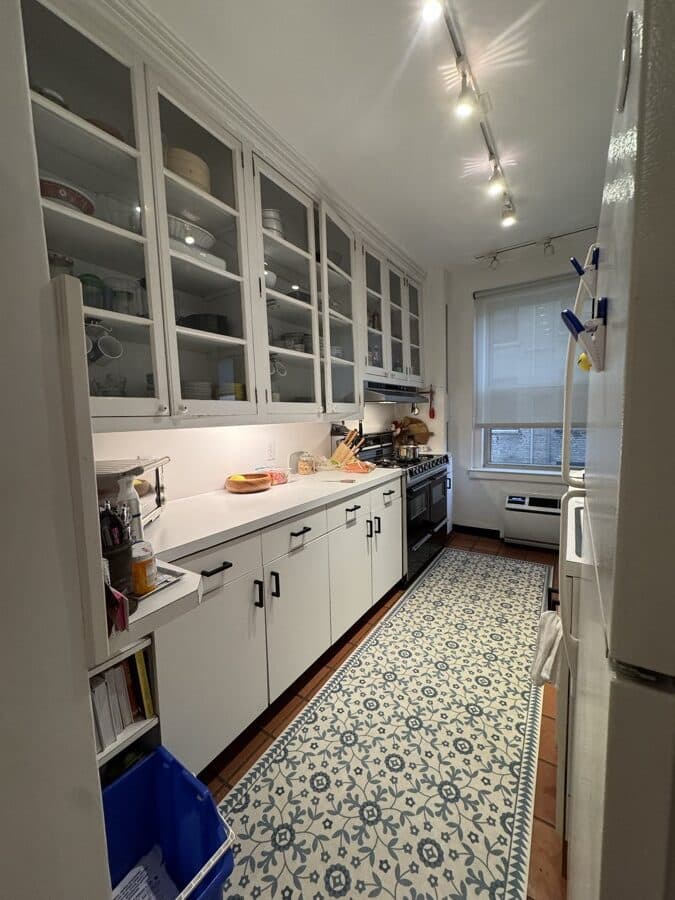 Before renovation: the original galley kitchen with white cabinets, glass-fronted uppers, ceiling track lighting and a pale patterned lino floor