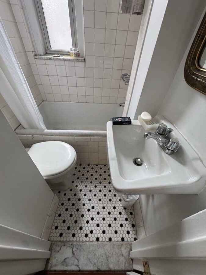 Before renovation: the office bathroom with white square wall tile, a wall-mounted sink with exposed trap and separate taps, and a black and white mosaic floor