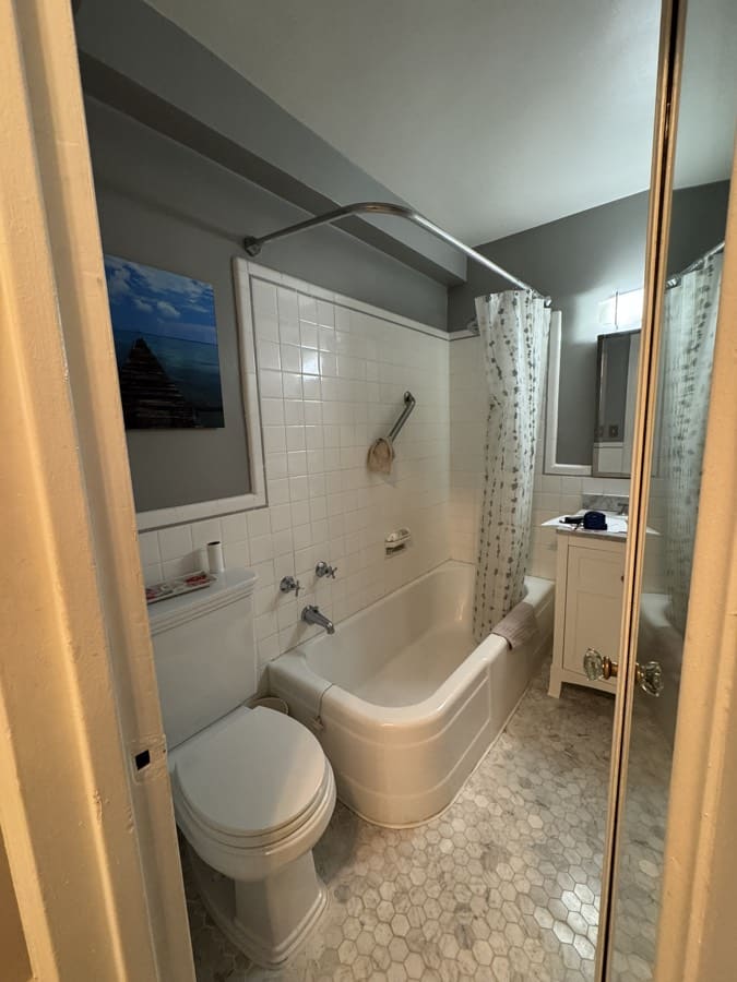Before renovation: the original cast-iron bathtub with a curved shower curtain rod, white square wall tile to half height and a 24 inch vanity