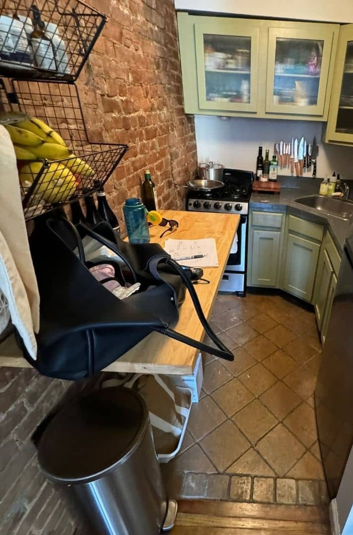 Before renovation: the original galley kitchen at 39 West 83rd Street with olive green glass-front cabinets, a dark laminate countertop, a freestanding gas range and terracotta floor tile, with the exposed brick wall at left