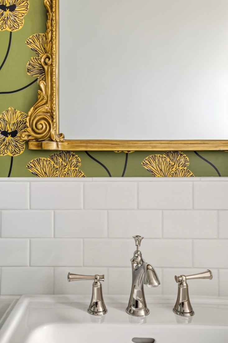 Finished bathroom detail: the Anthropologie Gleaming Primrose gold mirror against Mitchell Black Flourish floral wallpaper above Daltile Arctic White subway tile, with a Brizo Baliza polished nickel widespread faucet